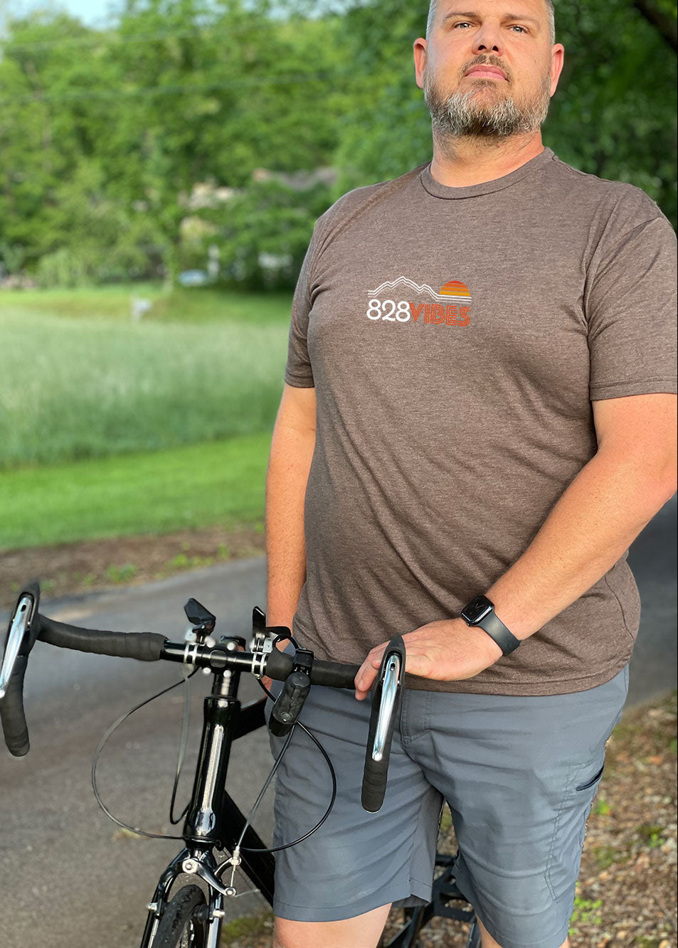 A person standing next to a bicycle, wearing a brown short-sleeve t-shirt with the '828Vibes' logo on it.