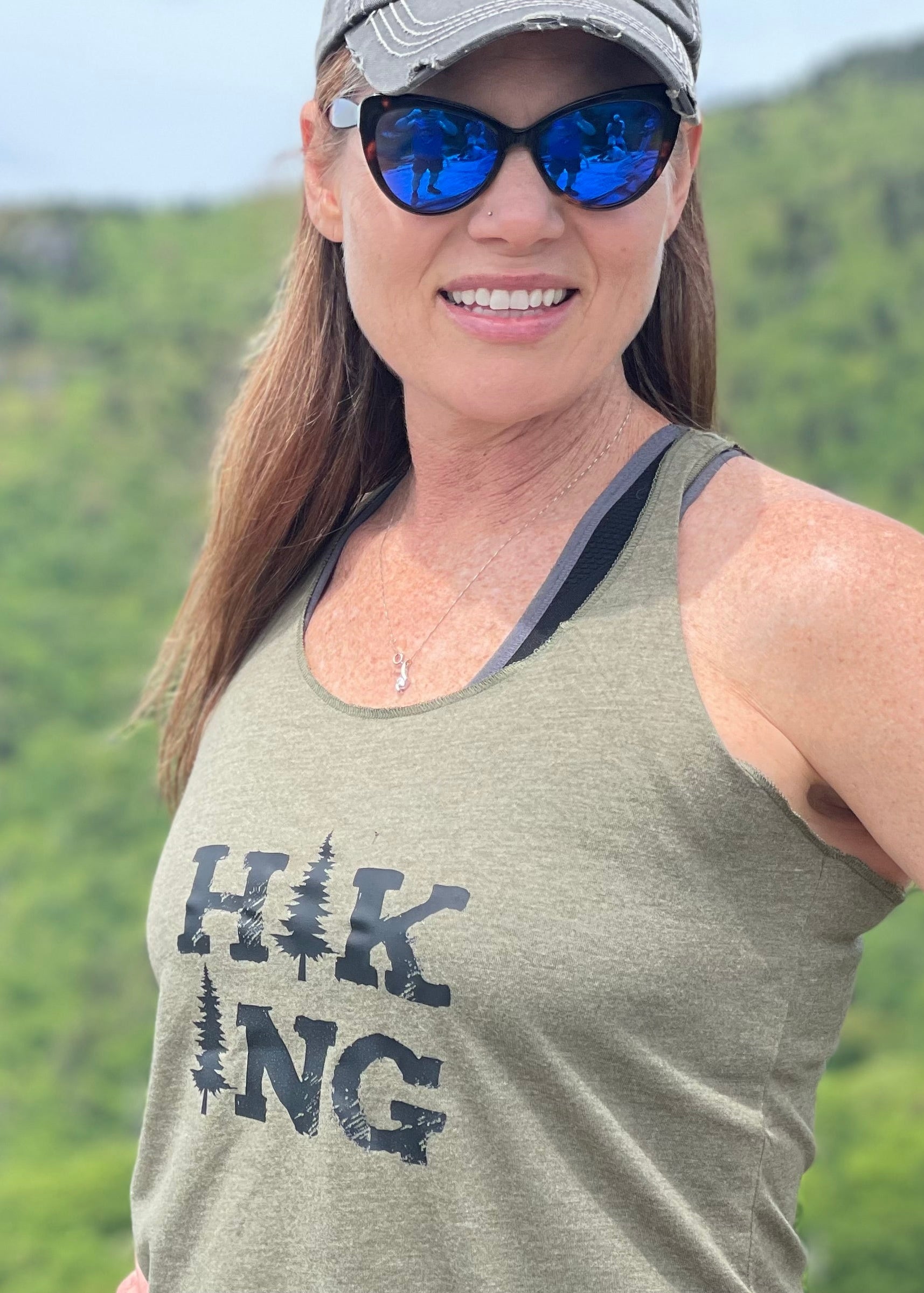 Woman wearing a green 'Hiking' tank top with sunglasses and a cap in a natural setting.