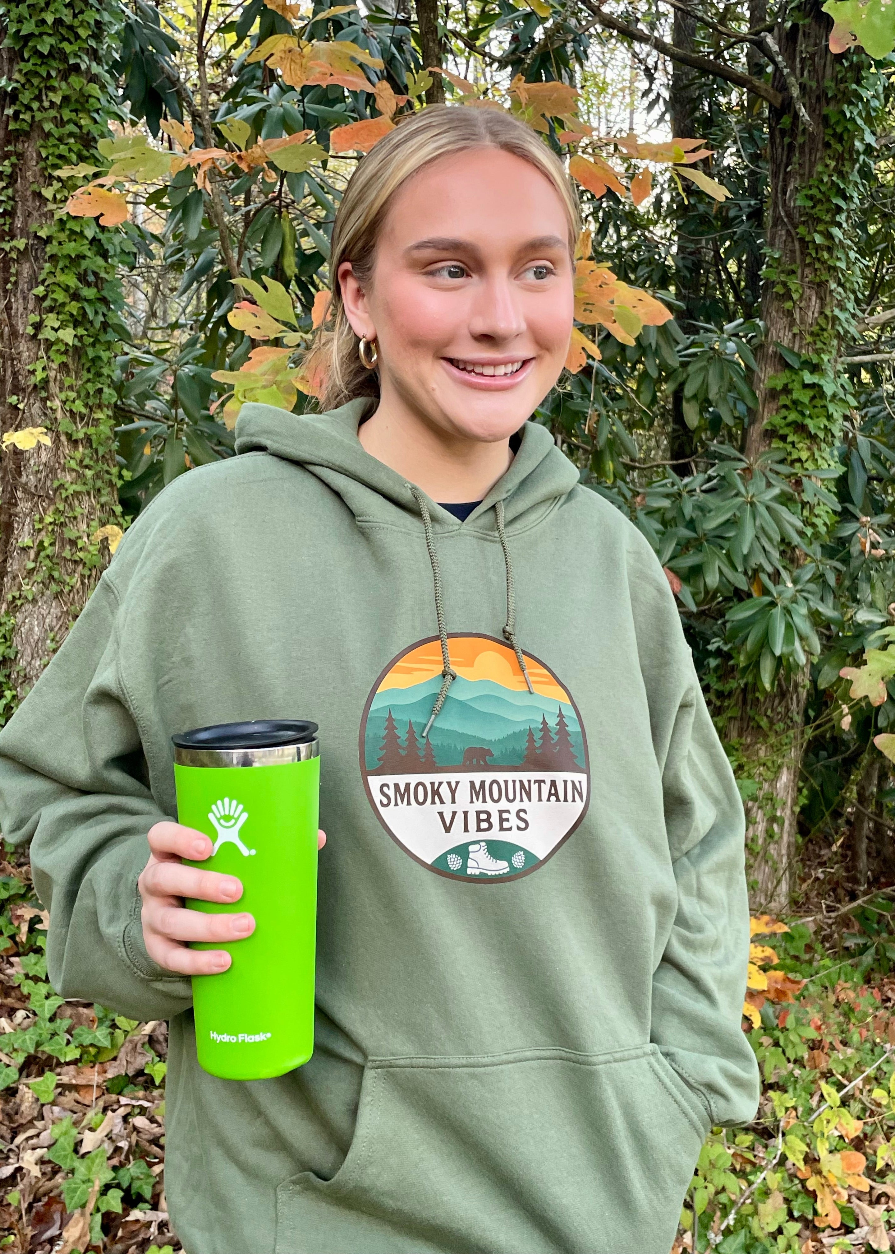 Person wearing a green hoodie with 'Smoky Mountain Vibes' design, holding a green tumbler in a forest setting.