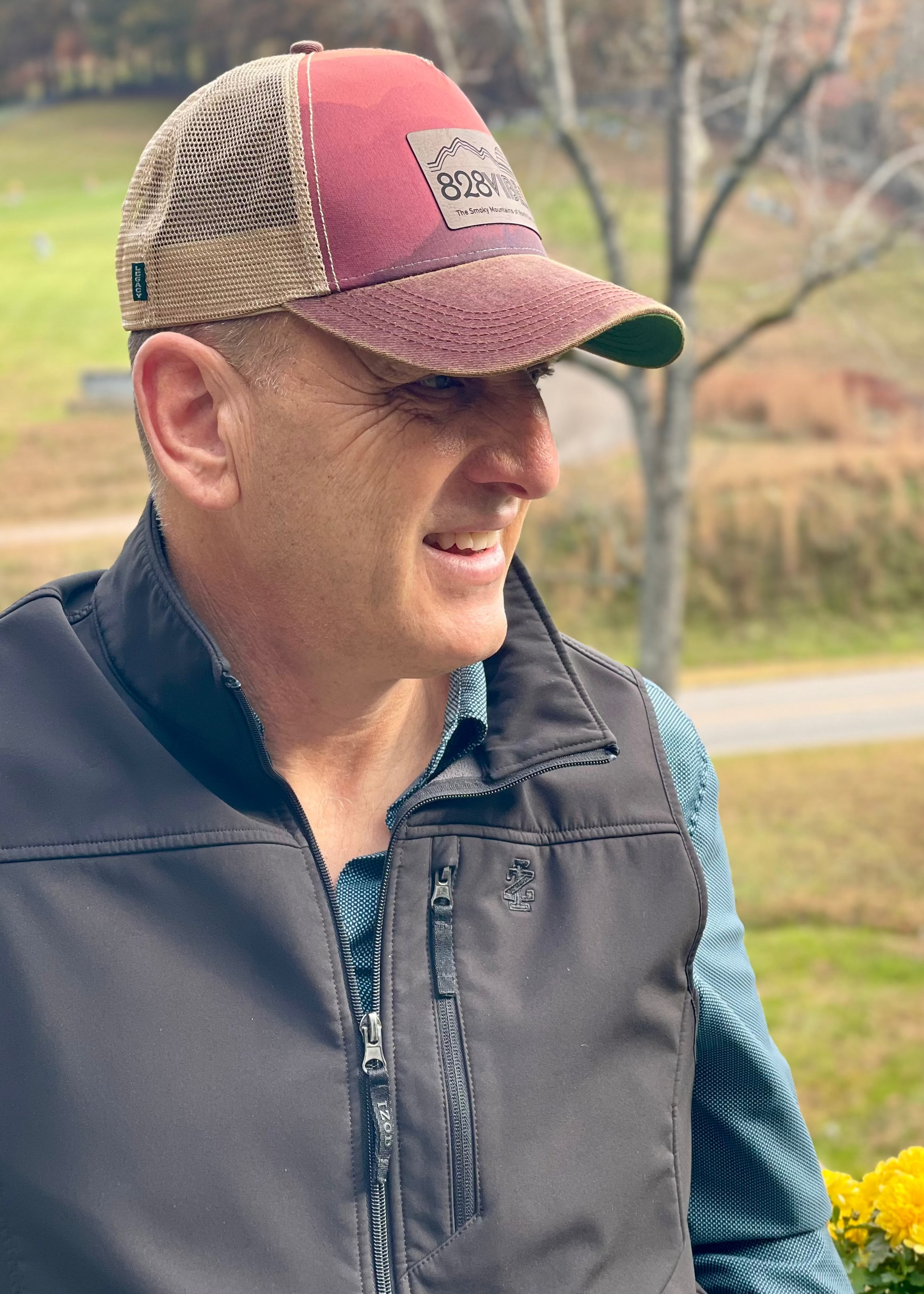 Man wearing a red and beige cap with a logo, standing outdoors in a park-like setting.