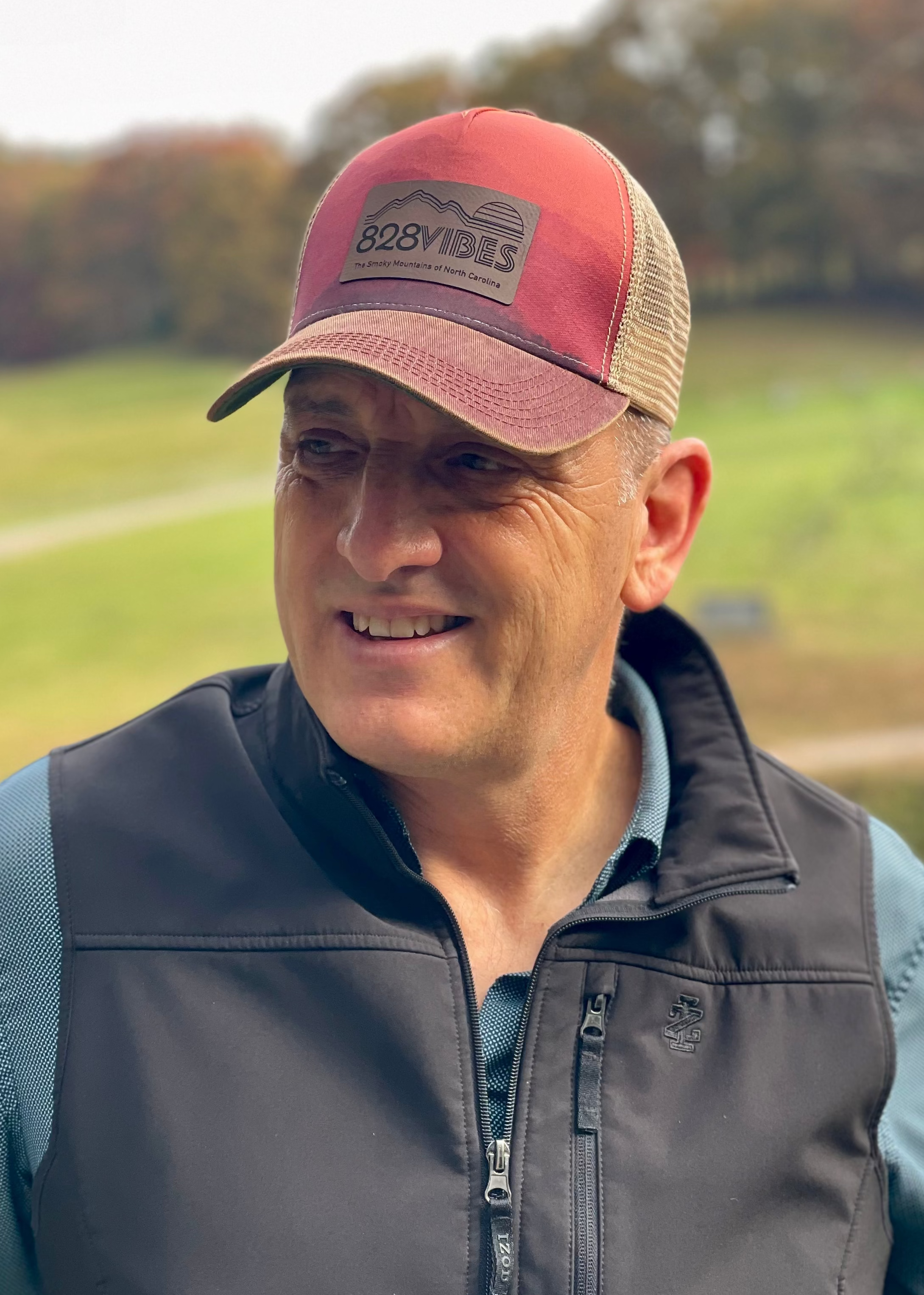 Man wearing a red cap and dark vest outdoors with greenery in the background