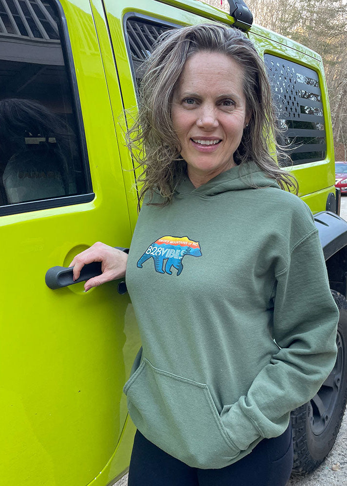 A woman standing beside a green Jeep, wearing a hoodie with a black bear graphic and the text '828 Vibes'. The hoodie is black, and the woman appears to be smiling.
