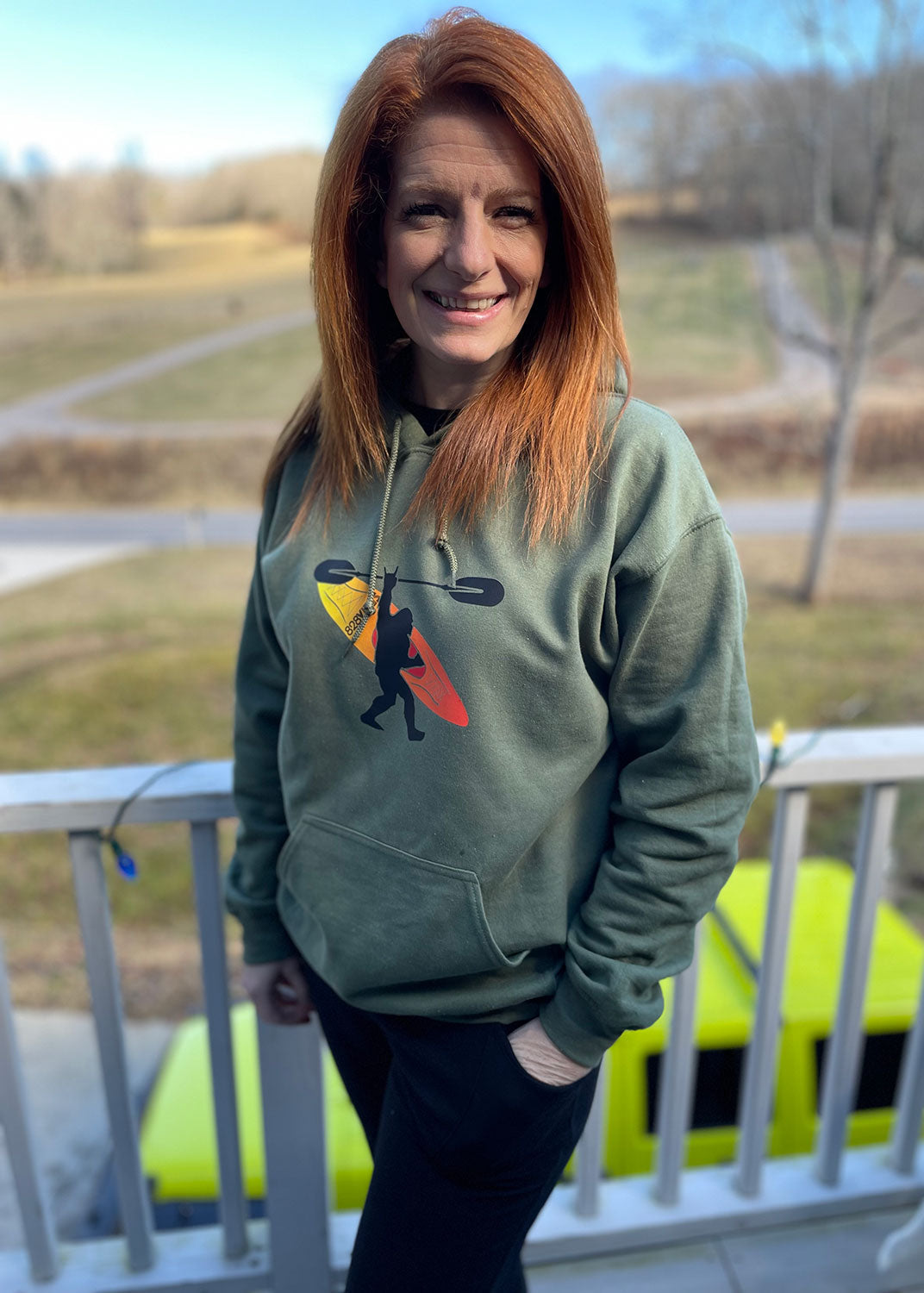 A person standing on a balcony wearing a green hoodie with a graphic of Bigfoot paddling a kayak in the Smoky Mountains.