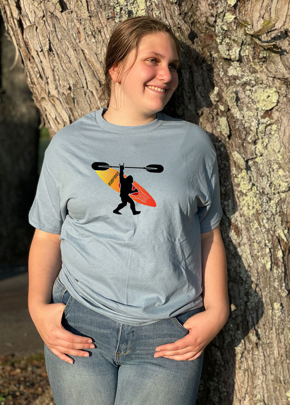 A person standing by a tree, wearing a short-sleeved T-shirt with a graphic of Bigfoot and a kayak on it.