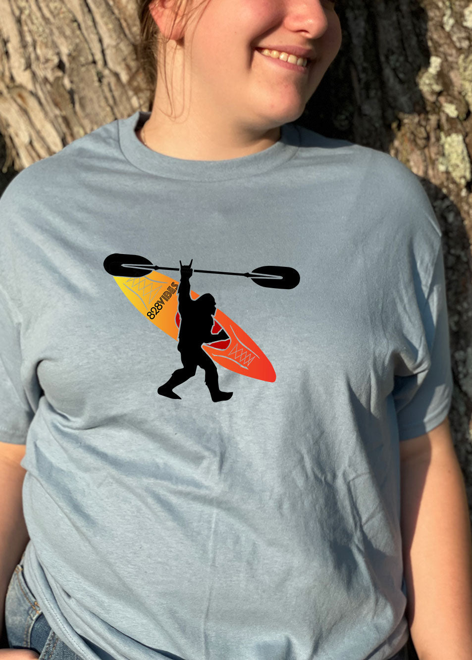 A person standing by a tree, wearing a short-sleeved T-shirt with a graphic of Bigfoot and a kayak on it.
