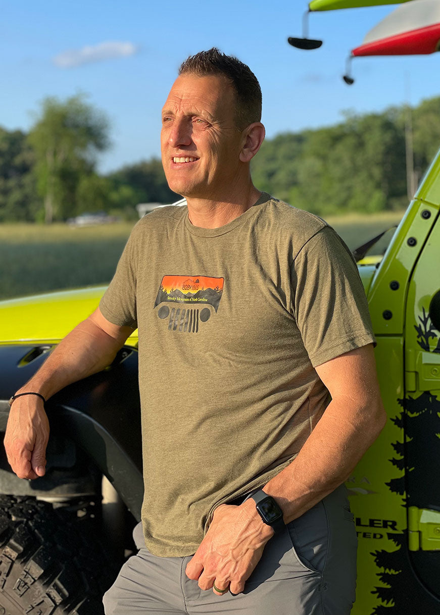 Man wearing a heather military green t-shirt with a Jeep logo, standing next to a yellow vehicle with kayaks on top.