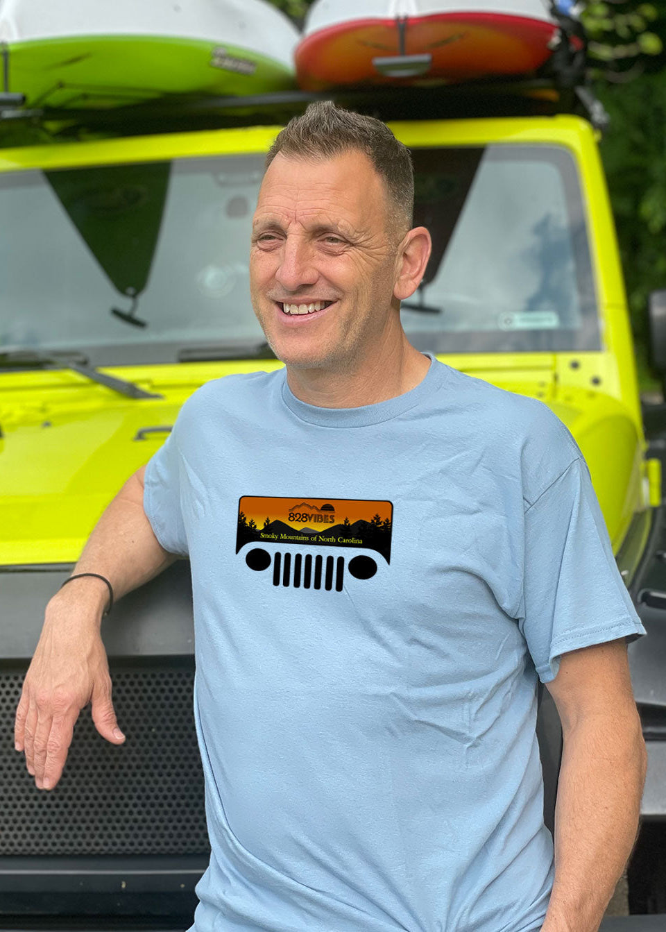 Man wearing a light blue t-shirt with a Jeep logo, standing next to a yellow vehicle with kayaks on top.