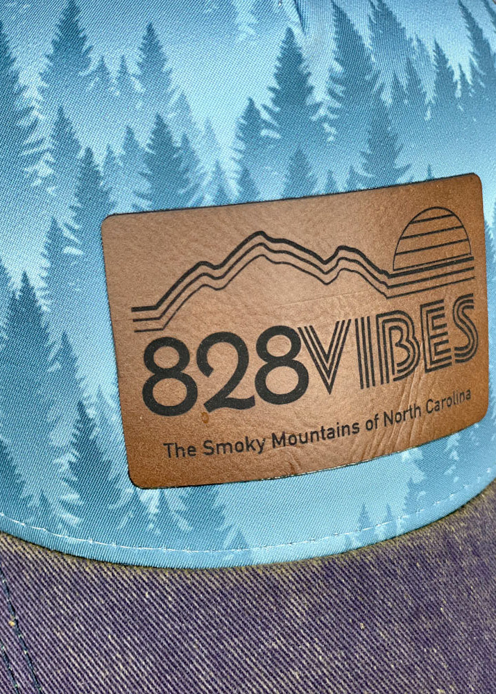 Cap with '828 Vibes' logo and mountain design against a blue background with tree pattern