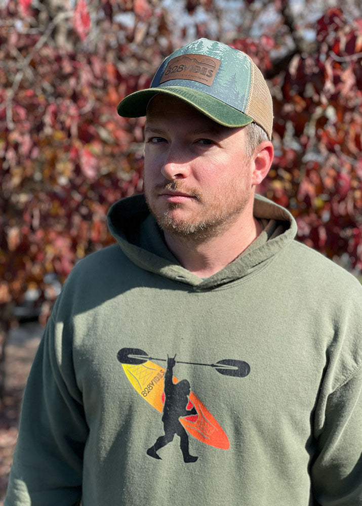 Man wearing a green hoodie with a paddle graphic and cap, standing in front of a wall of red and orange flags.