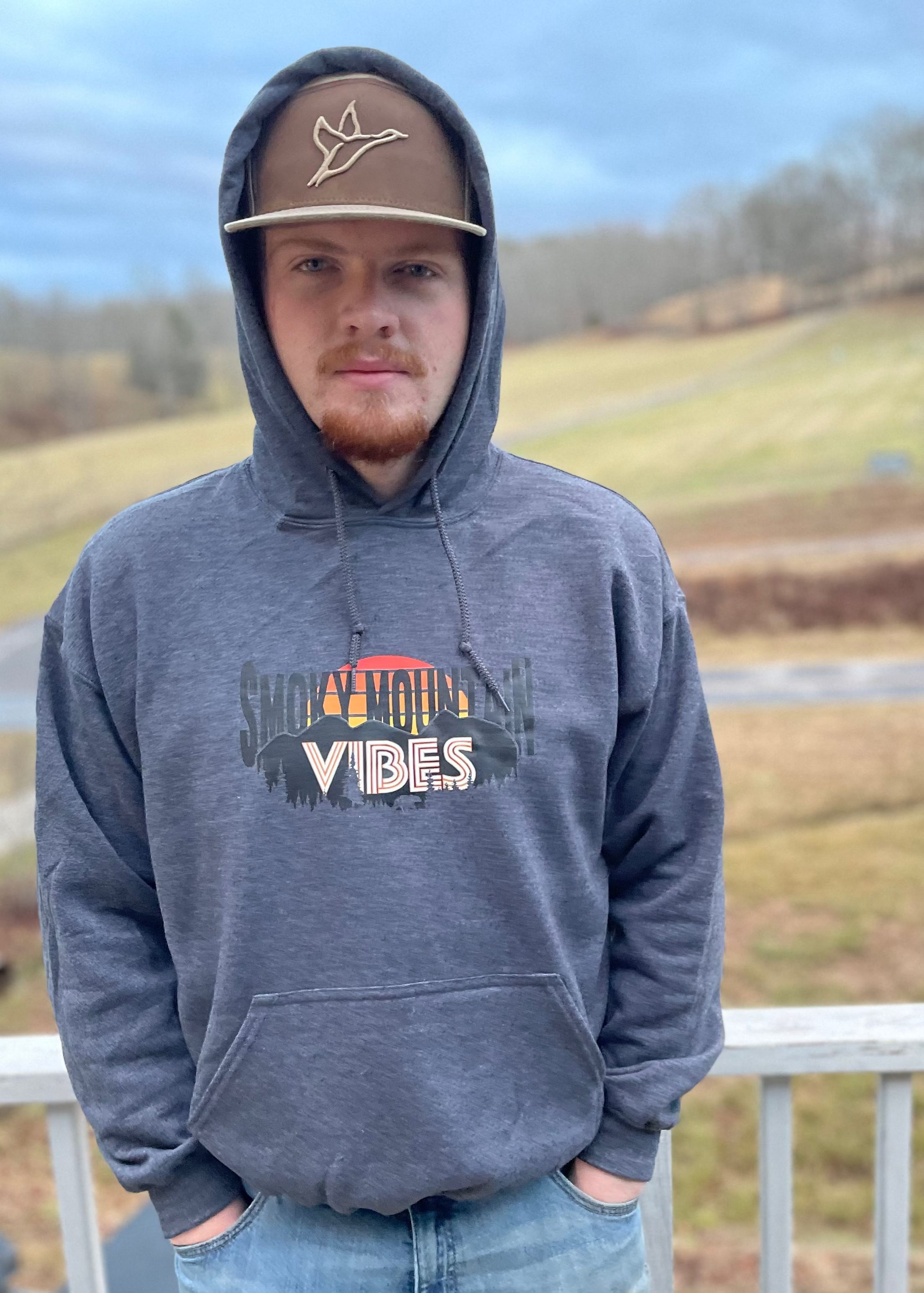 A man wearing a gray hoodie with 'Smoky Mountains Vibes' text and a graphic design on the front, standing outdoors.