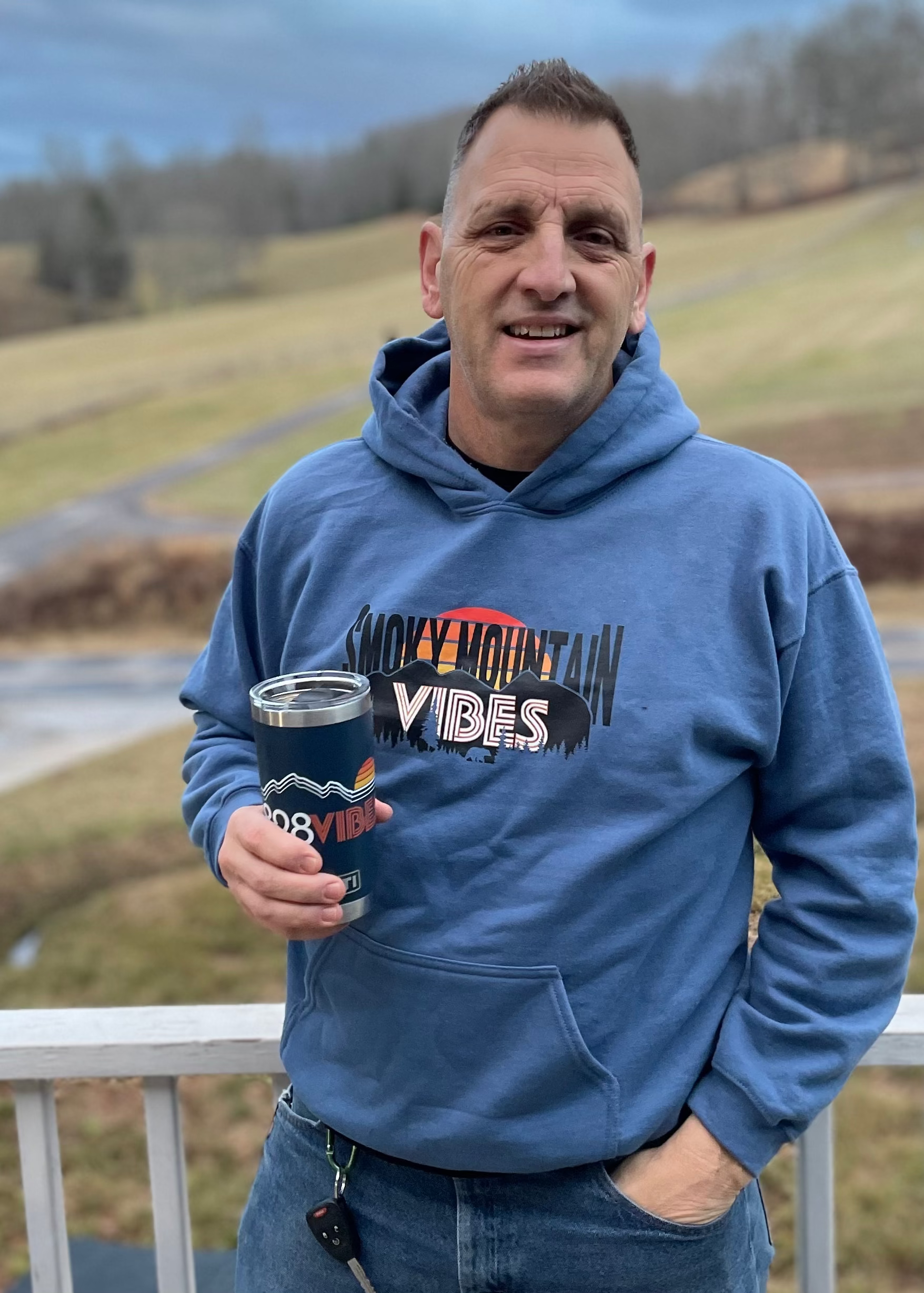 Man wearing a blue hoodie with 'Smoky Mountain Vibes' text, holding a cup outdoors.