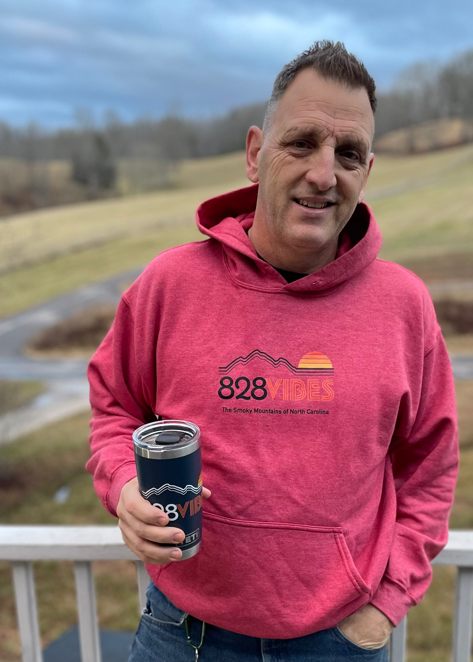 A man standing outdoors wearing a pink hoodie with the '828 Vibes' logo and a blue kangaroo pocket.