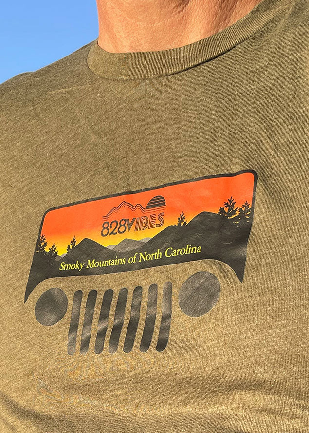 Man wearing a heather military green t-shirt with a Jeep logo, standing next to a yellow vehicle with kayaks on top.