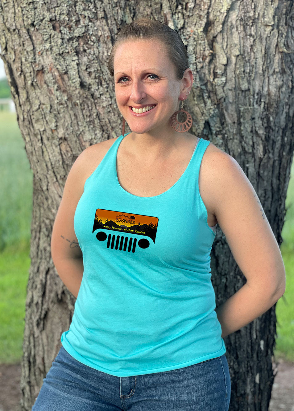 A woman standing by a tree, wearing a blue racerback tank top with a Jeep grill graphic.
